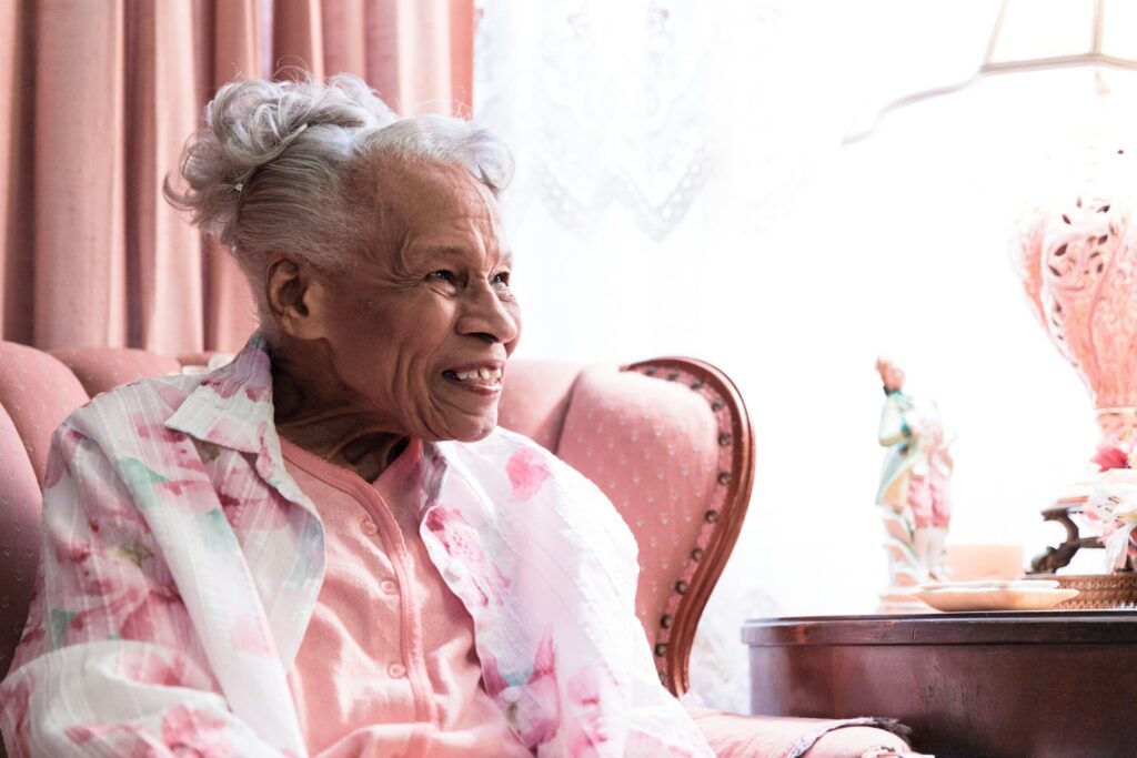 An older woman wearing pink sitting in a pink chair smiles