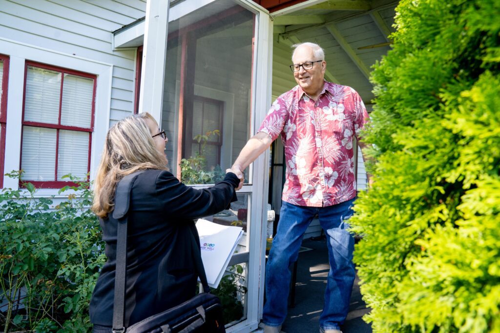An Age Well with a binder and bag member greets a man on his porch wearing a pink Hawaiian shirt