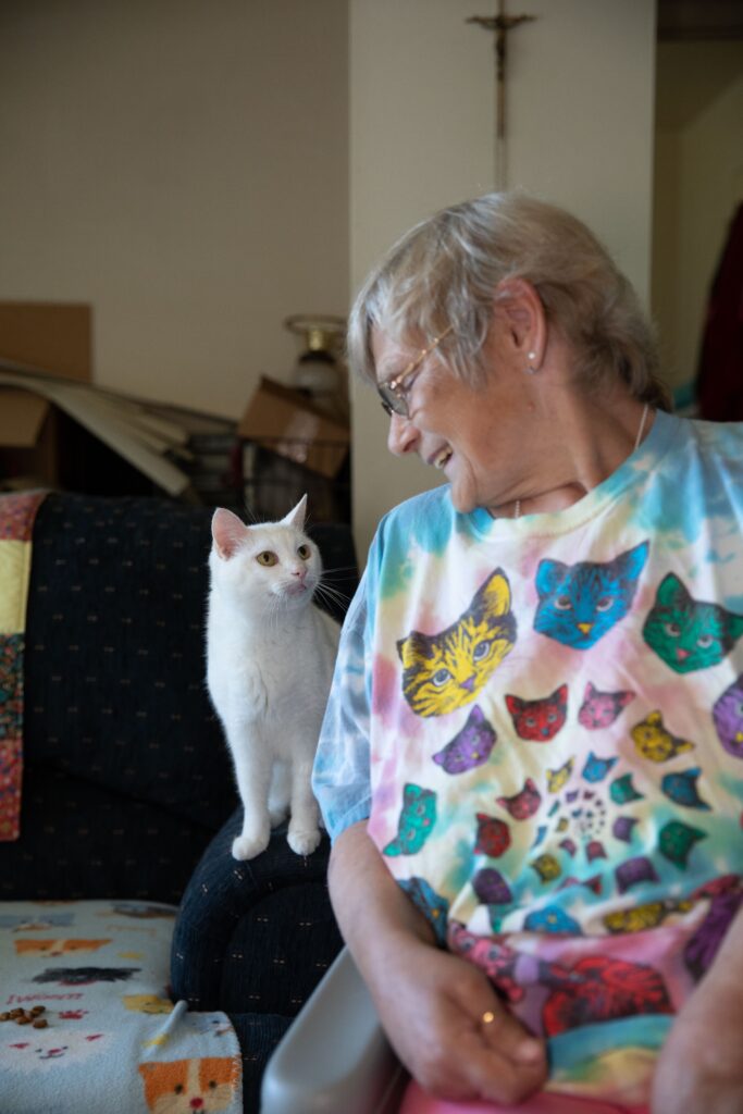 A sitting older woman smiles as she looks to her right to see her white cat looking back at her