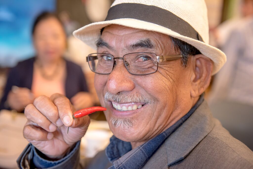 An older man wearing a white hat and glasses smiles as he's about to eat a small red pepper