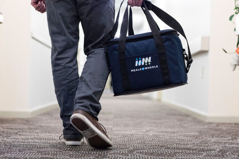 A person with gray pants, walking down a hallway carrying a blue food cooler with the Meals for Wheel logo embroidered on it