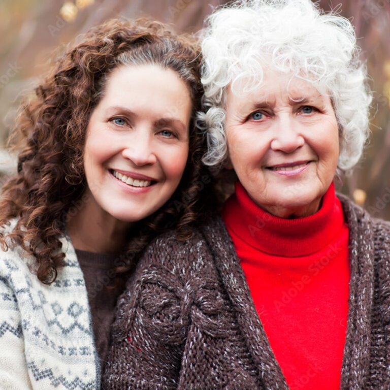 Photograph of two women smiling towards the camera. The woman on the left has brown hair and is a family caregiver for the senior woman on the right with white hair.