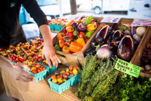 Photograph of produce on a table at a farmers market including cherry tomatoes, fresh herbs, large eggplant. A light skinned hand is pictured on the left reaching for a pint of cherry tomatoes.